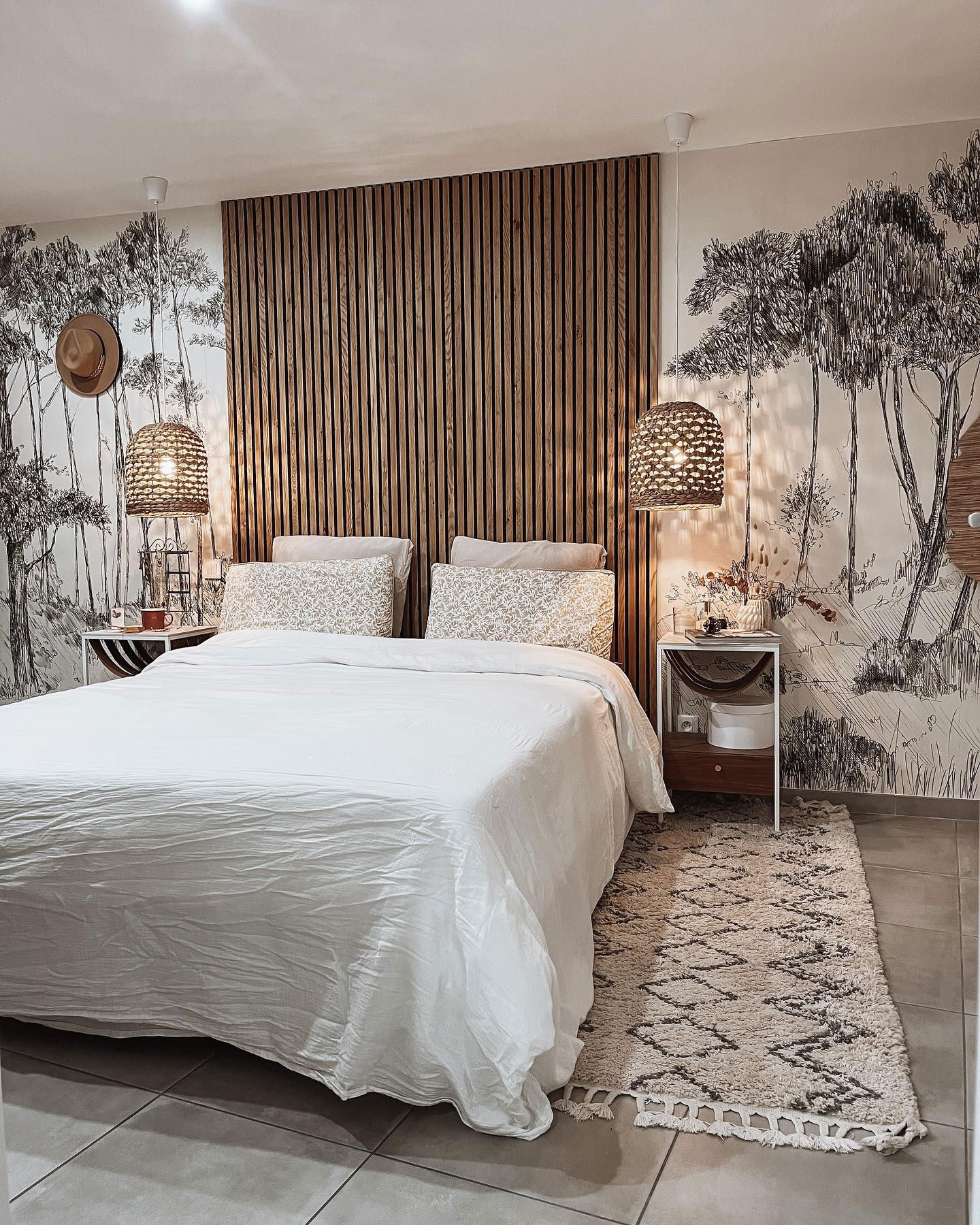 Bedroom with white bedding, wood slat headboard, and forest mural. Woven pendant lights and patterned rug.
