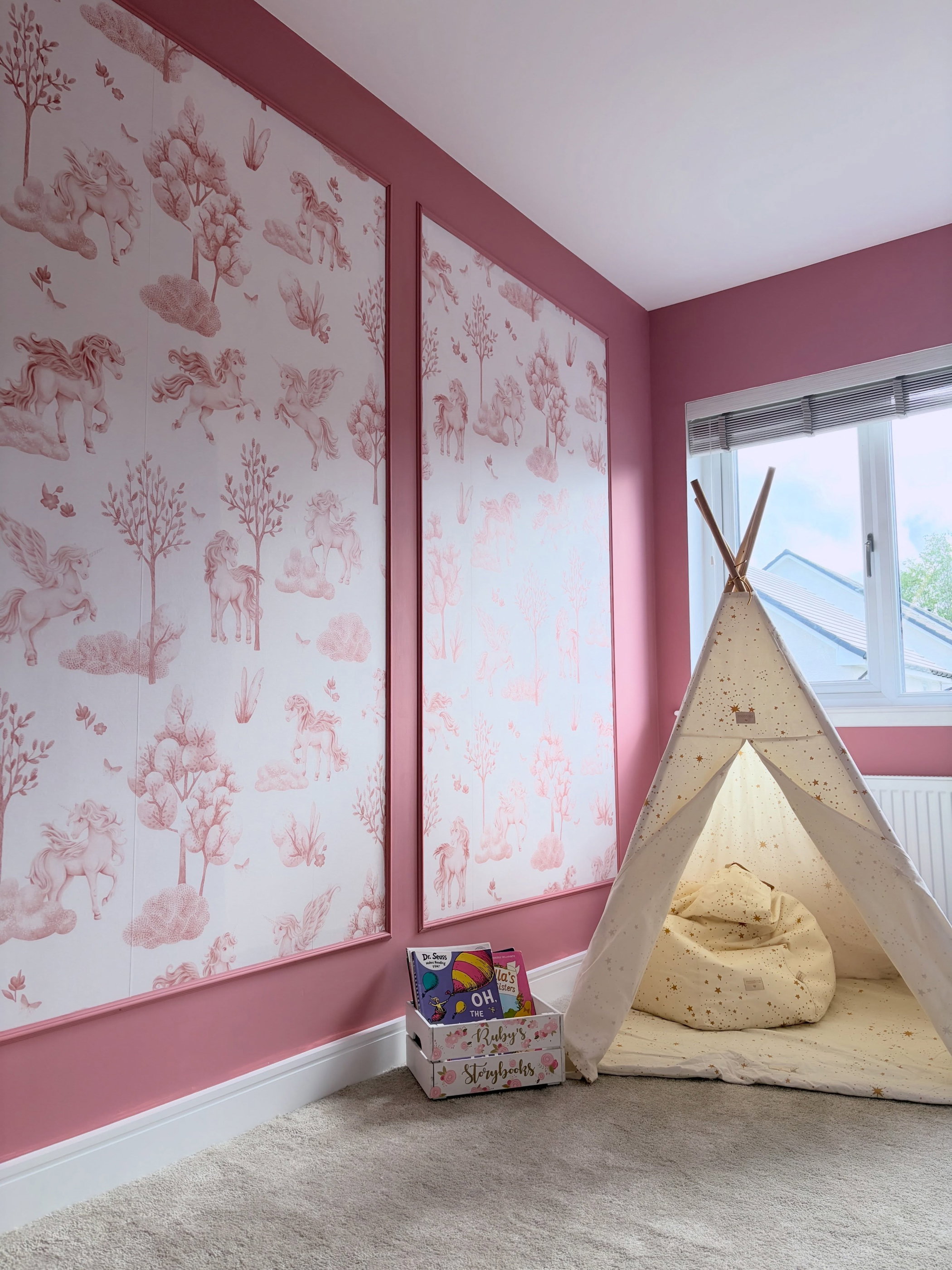 Ett barnrum med rosa väggar, två fondtapeter med rosa enhörningar och pegaser, ett krämfärgat tipi-tält med en saccosäck och en boklåda på grå heltäckningsmatta.
