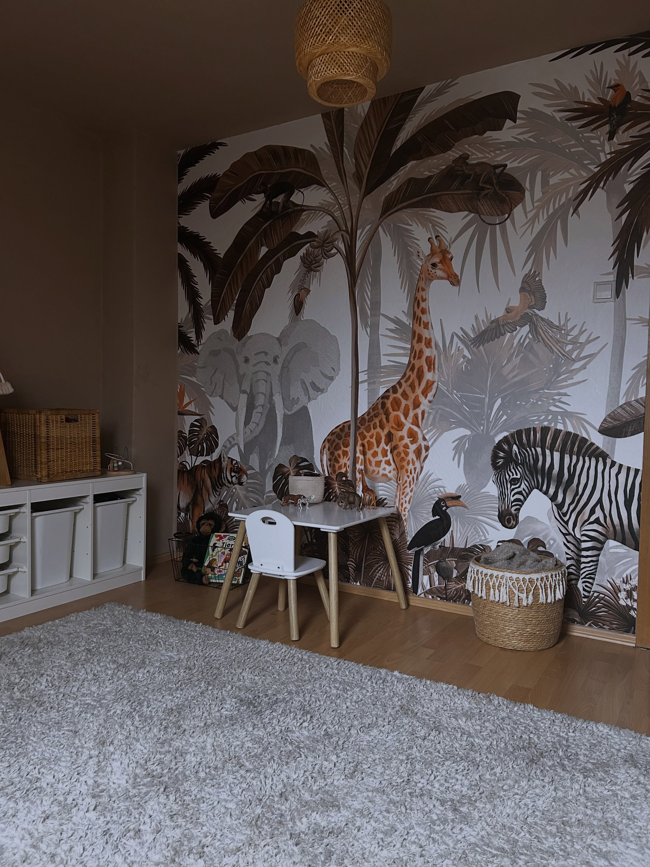 Kinderkamer met een jungle-muurschildering met een olifant, giraf en zebra. Op de voorgrond een witte tafel en stoel, een opbergmeubel en een lichtgrijs hoogpolig vloerkleed.
