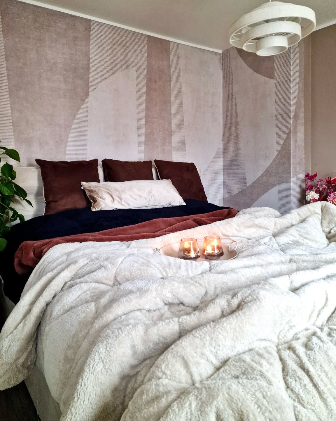 Un letto accogliente con una coperta soffice grigio chiaro, coperte blu scuro e ruggine, cuscini marroni e bianchi e due candele accese su un vassoio. Dietro il letto c'è una carta da parati astratta geometrica.