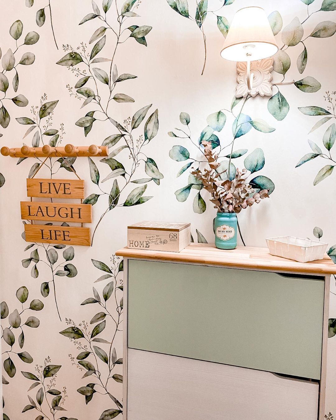 Interior with leaf-patterned wallpaper, wall lamp, and two-tone green and white cabinet. "Live Laugh Life" sign on wall.