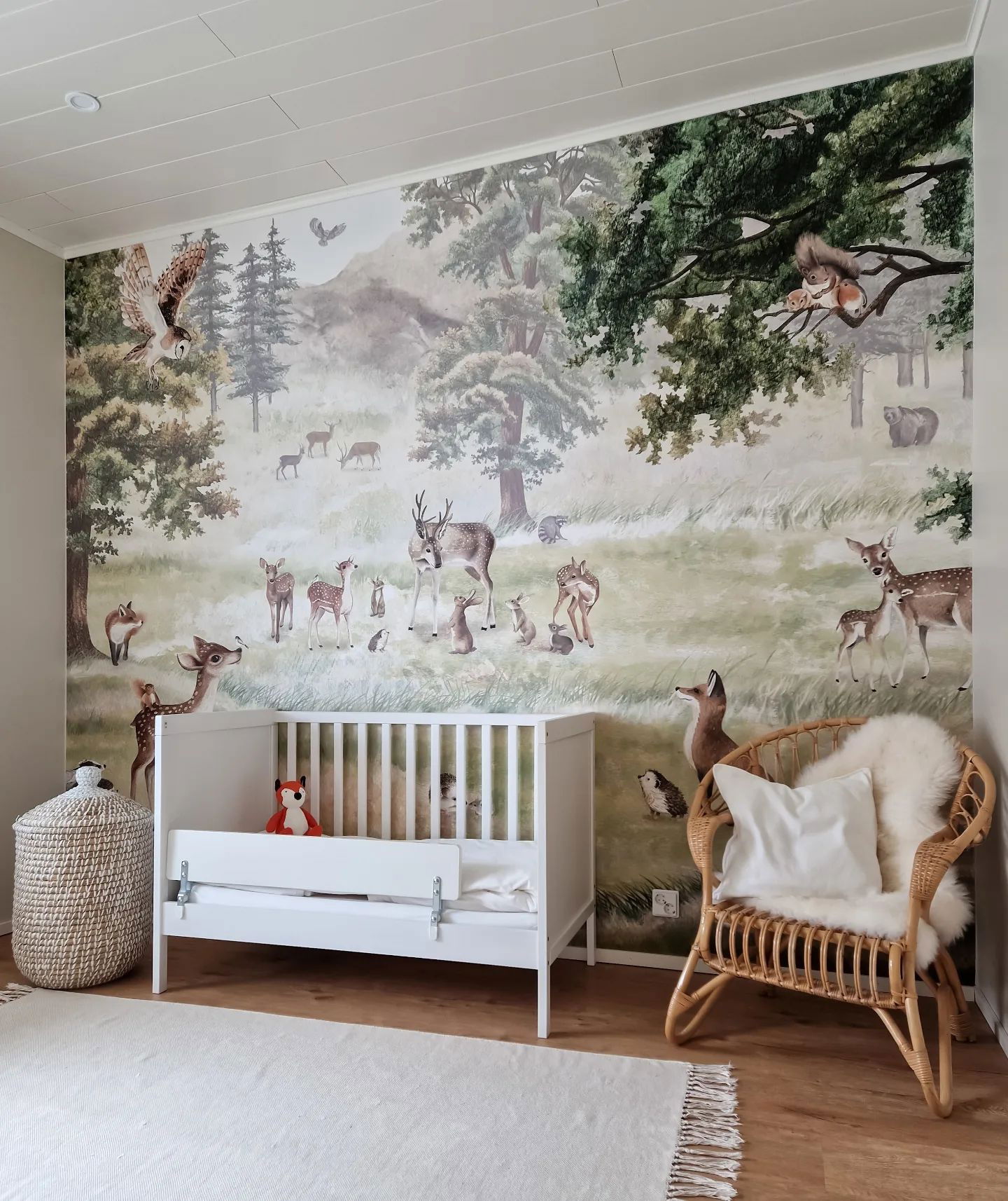 Chambre de bébé avec une peinture murale de forêt, un berceau blanc et une chaise en osier. La peinture murale représente des cerfs, des renards, des lapins et d'autres créatures des bois.