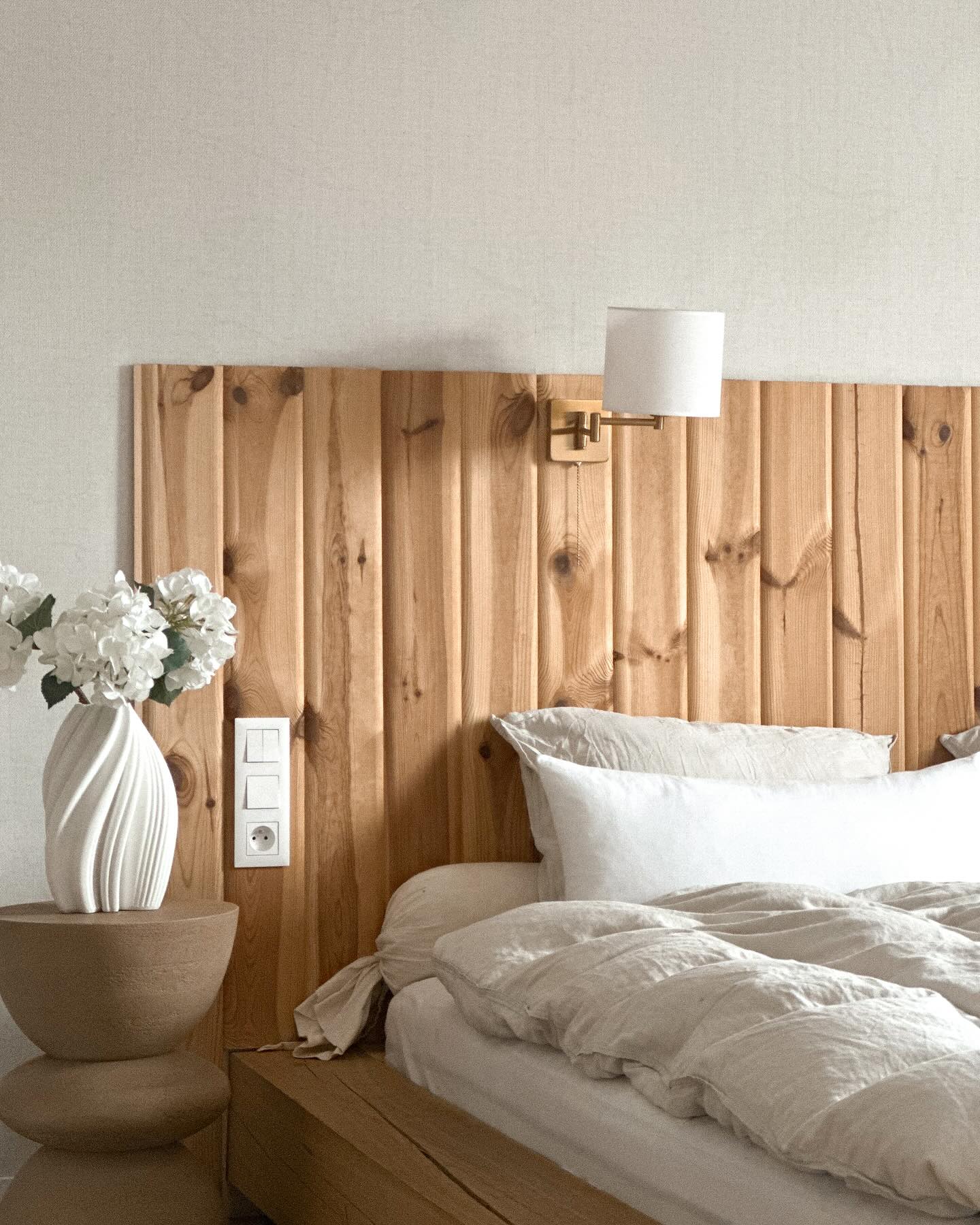 Soverom med sengegavl i tre, hvitt sengetøy og en vase med hvite blomster på et rundt sidebord.