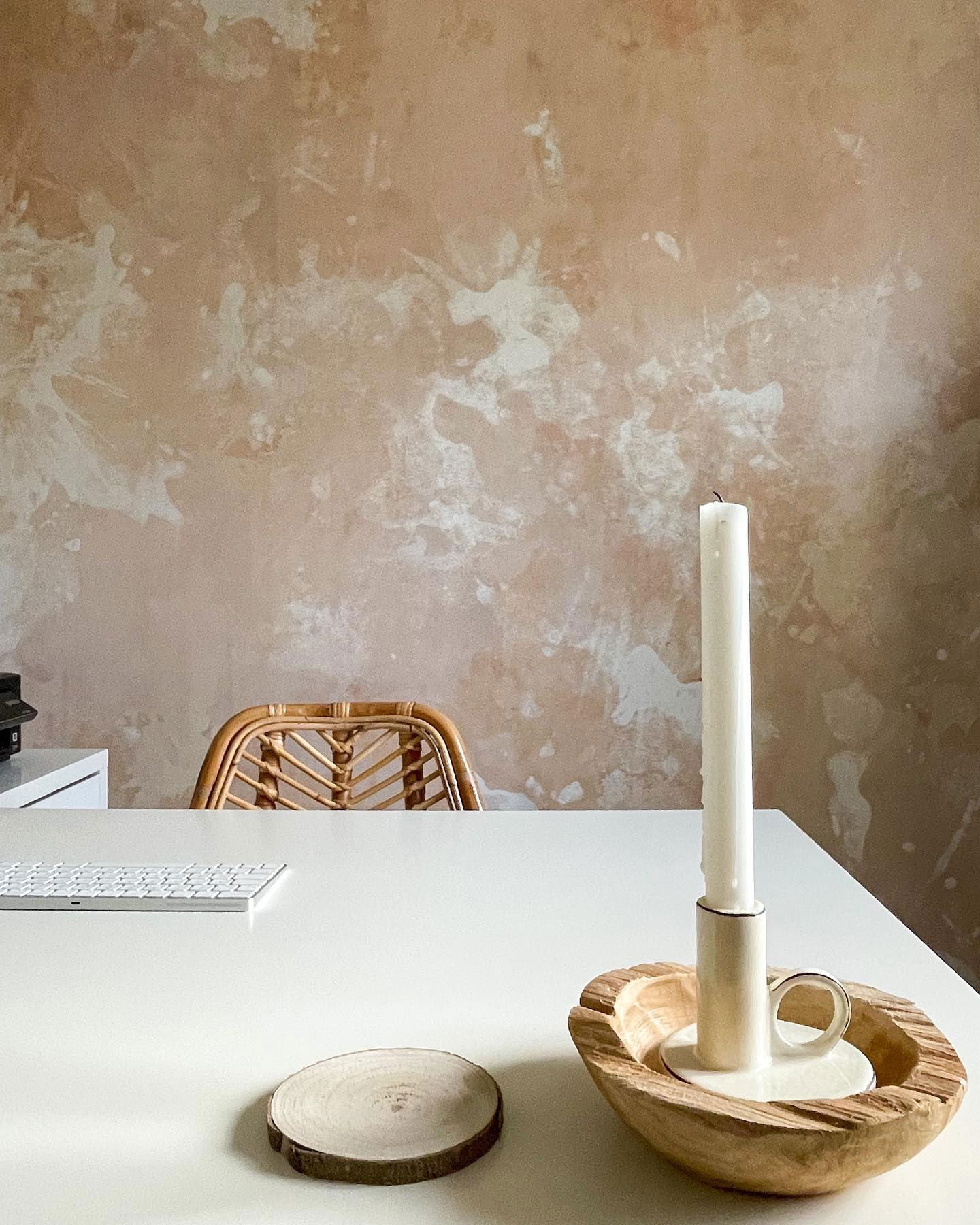 Minimalistischer Arbeitsbereich mit weißem Schreibtisch, Tastatur, Kerze in Holzhalter und Rattanstuhl vor einer strukturierten Wand.