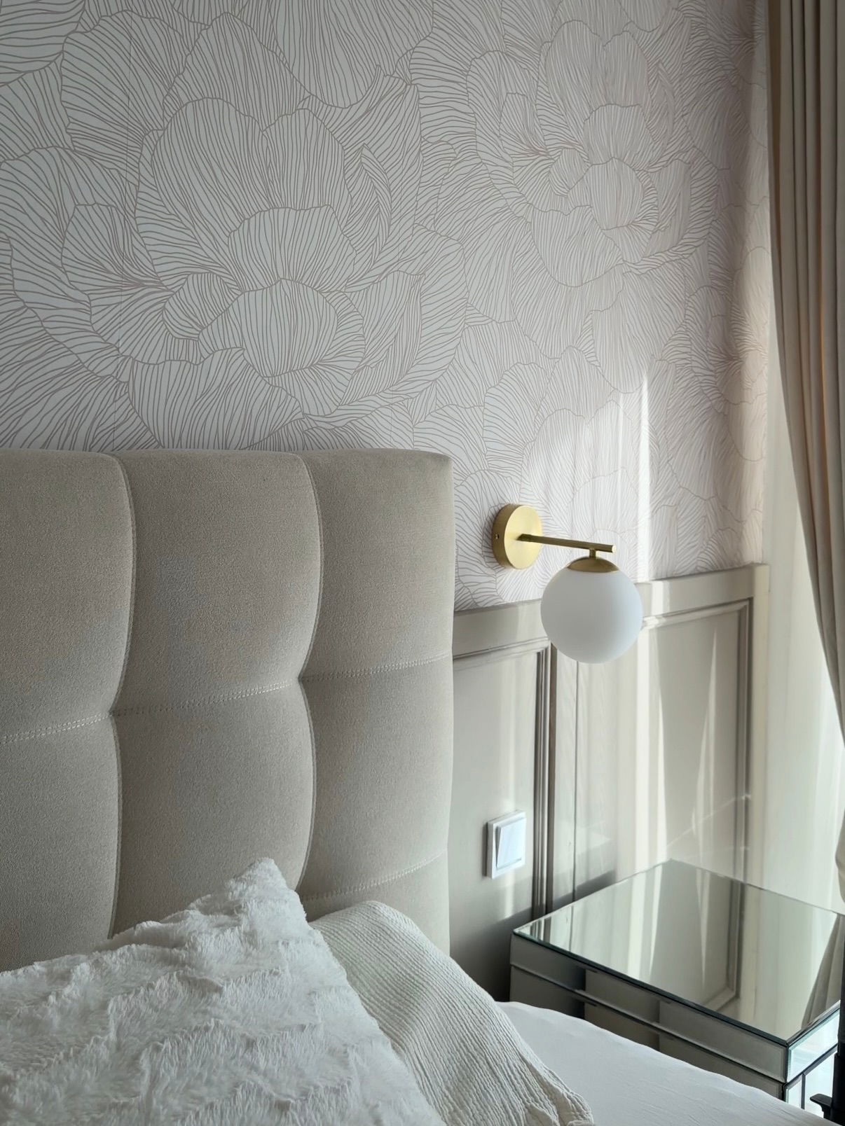 Tufted cream headboard with ornate wallpaper, gold sconce with white globe, and mirrored nightstand.