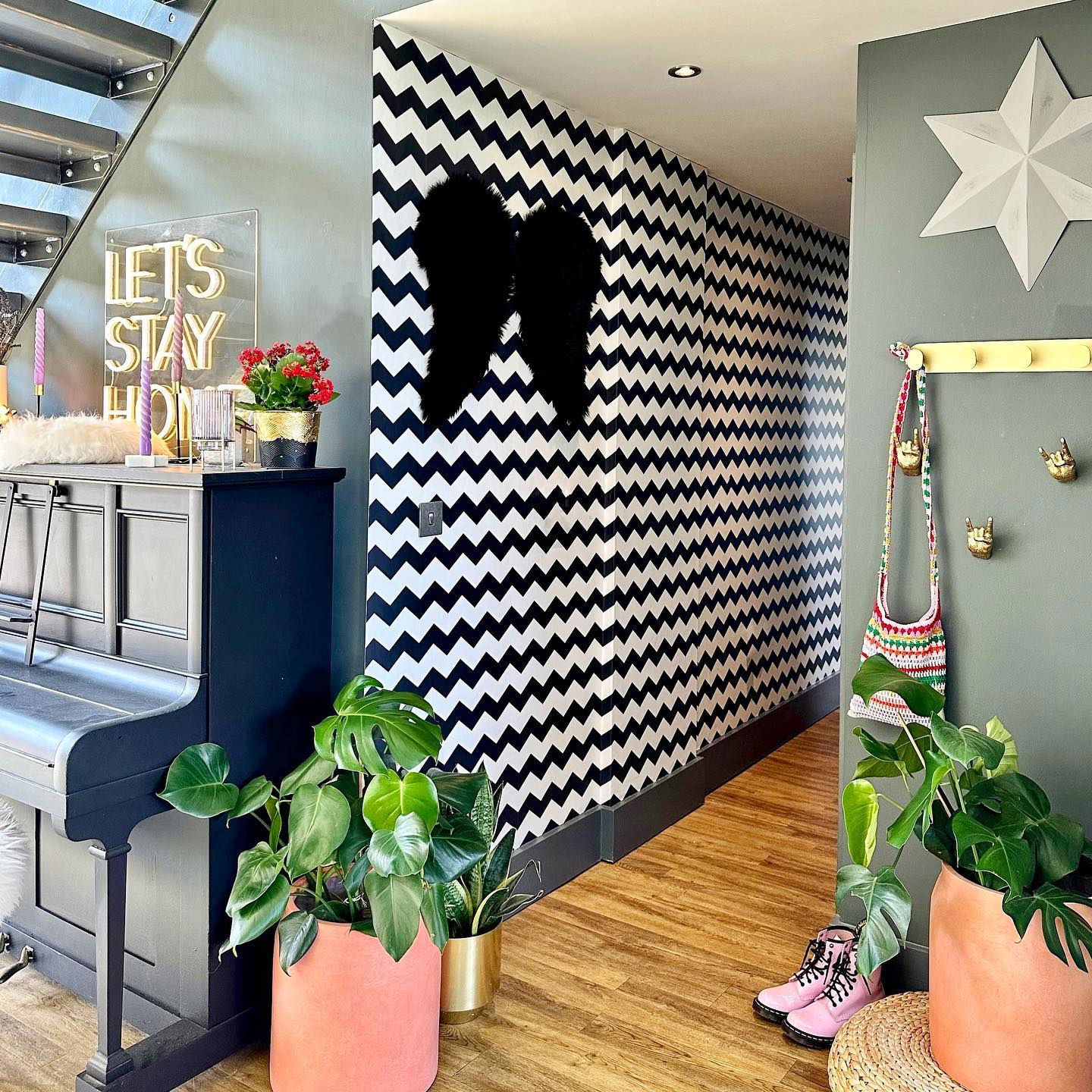 Eclectisch interieur met zwarte piano, chevronbehang en potplanten. Roze laarzen en een neonbord met de tekst "Let's Stay Home" voegen een speels element toe.