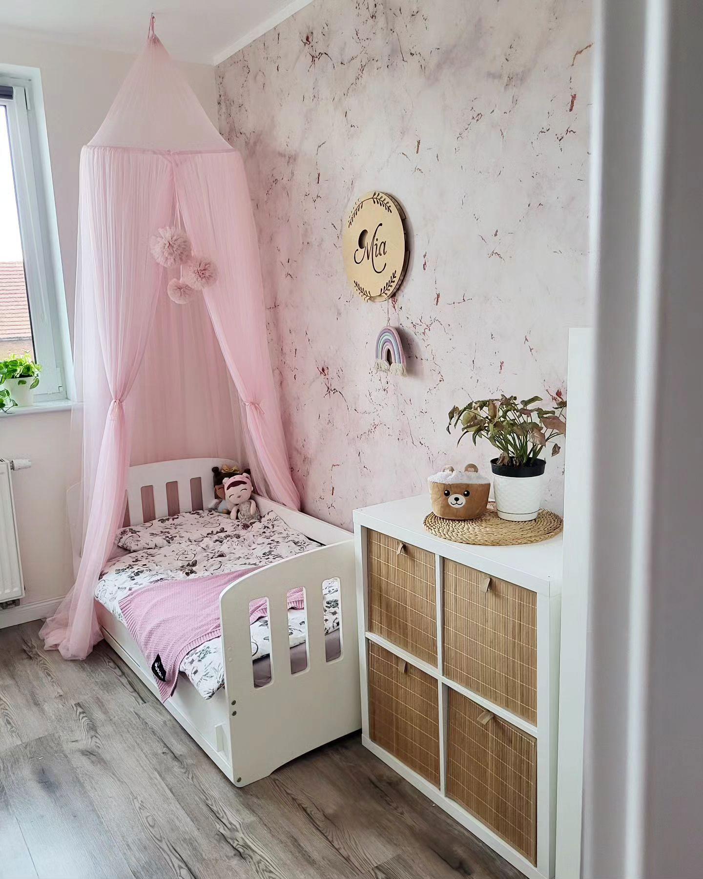 Habitación infantil con cama blanca bajo dosel rosa, unidad de almacenamiento blanca con cajones de mimbre y papel pintado de mármol rosa.