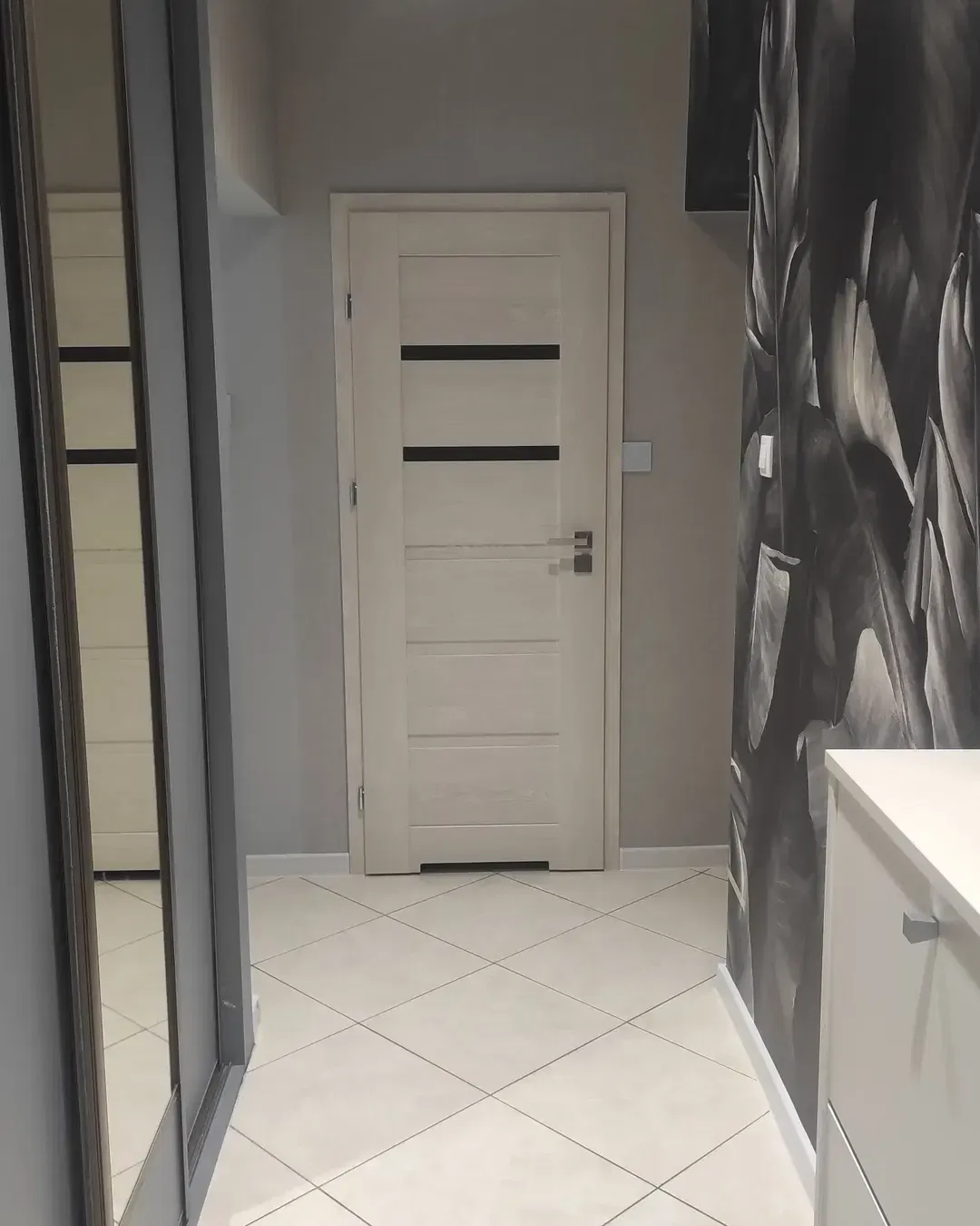Couloir avec porte blanche, murs gris et carrelage blanc. Un placard à miroir se trouve sur la gauche.