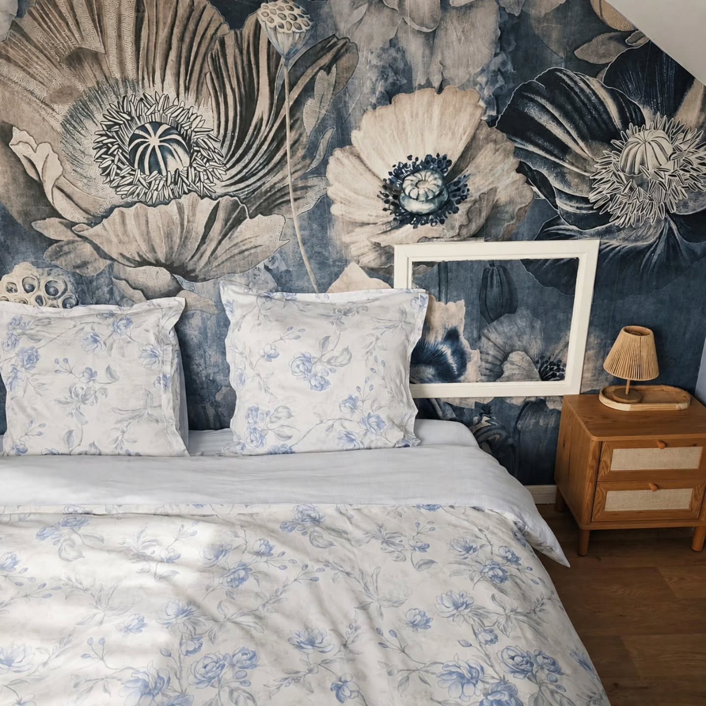 Bed with white and blue floral bedding against a wall with large dark blue and light gray floral wallpaper. A light wood nightstand with a woven lamp is on the right.