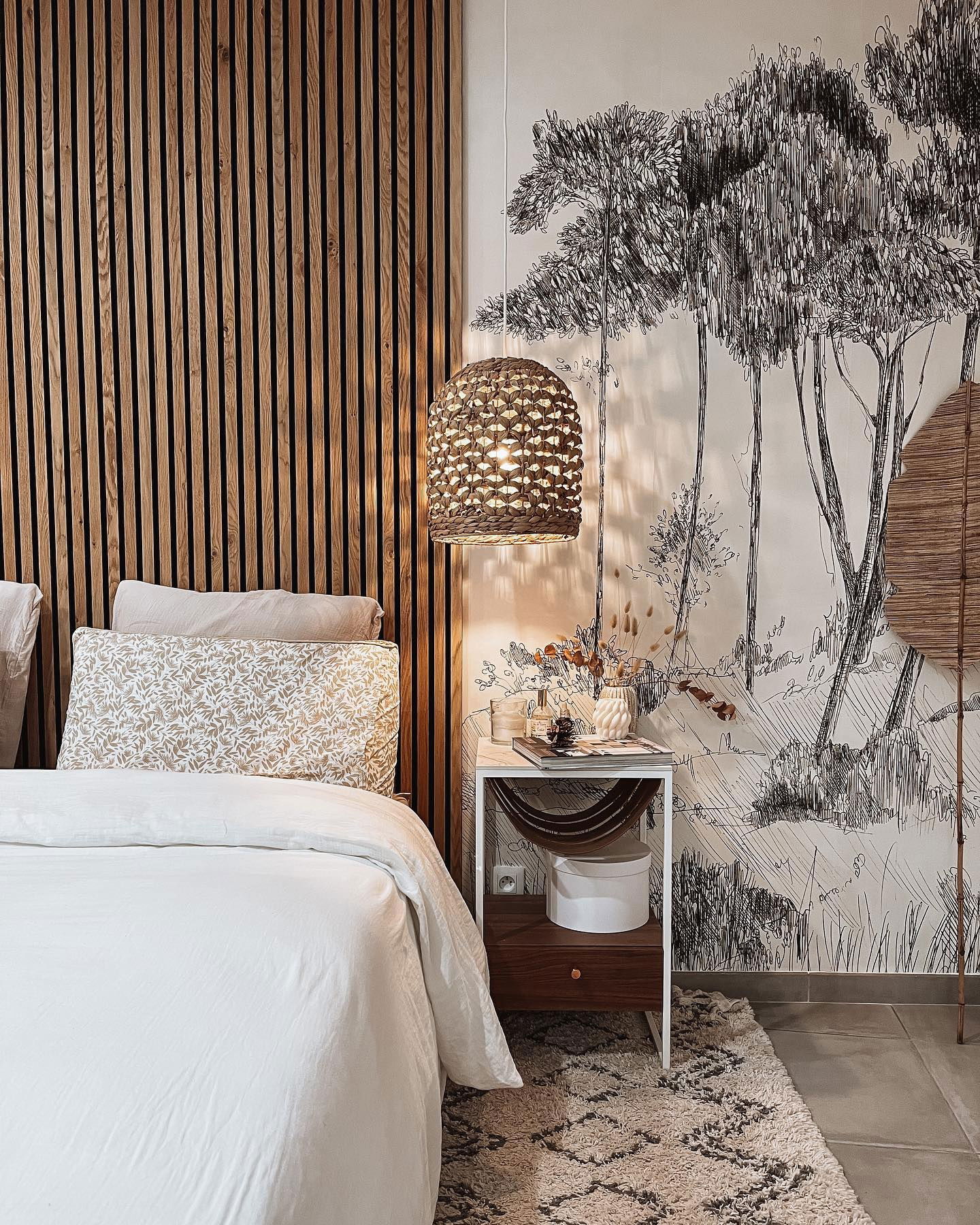 Schlafzimmer mit weißer Bettwäsche, Holzlatten-Kopfteil und Baumskizzen-Wandkunst. Rattan-Hängeleuchte und Nachttisch mit Dekor.