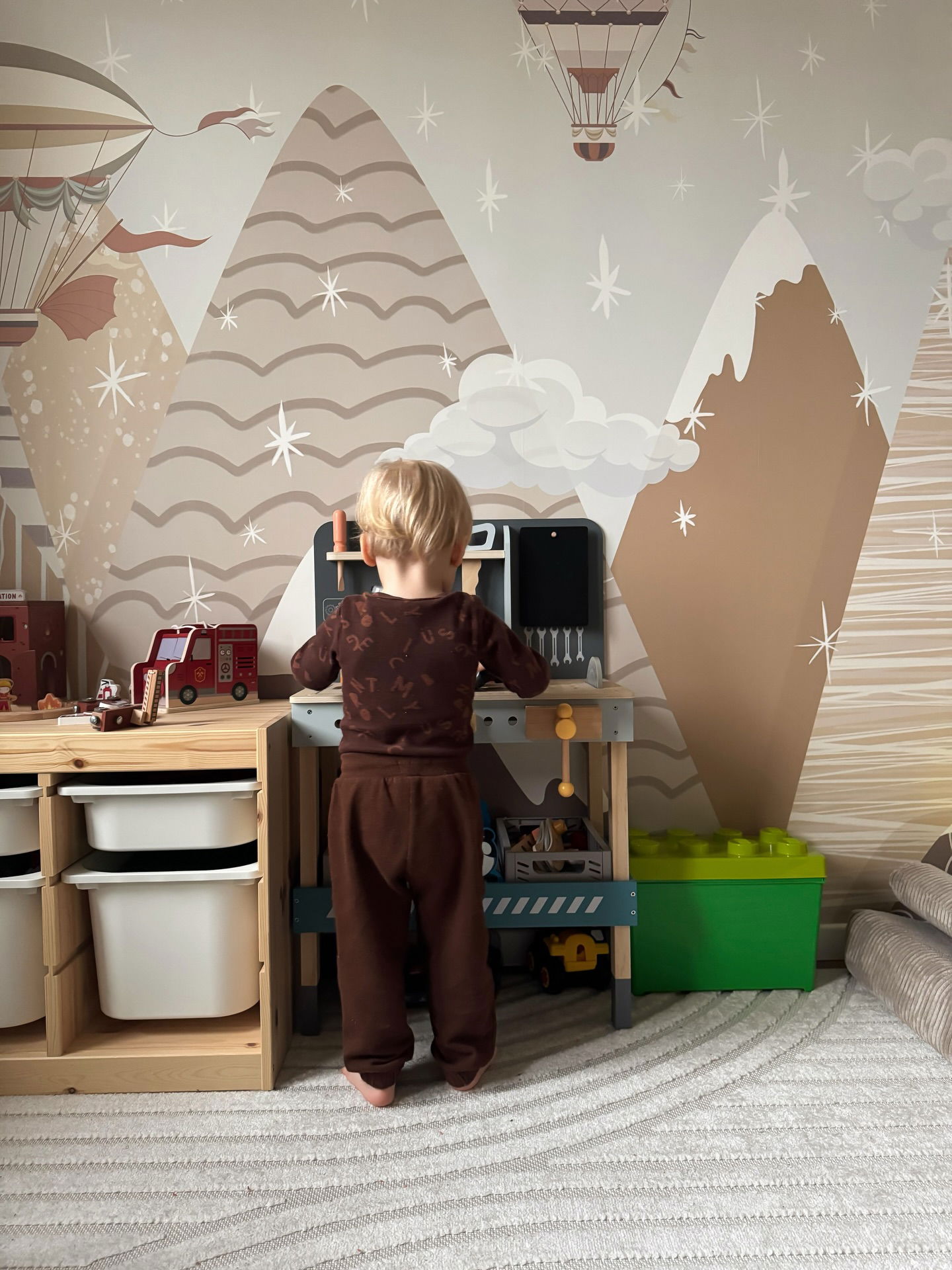 Blondes Kleinkind in braunem Schlafanzug spielt an einer Spielzeugwerkbank in einem Raum mit tapeten im Bergdesign.