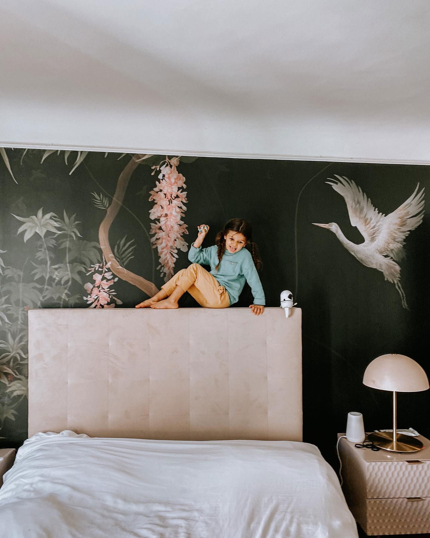 A girl sits on a bed's headboard in front of a dark floral mural.