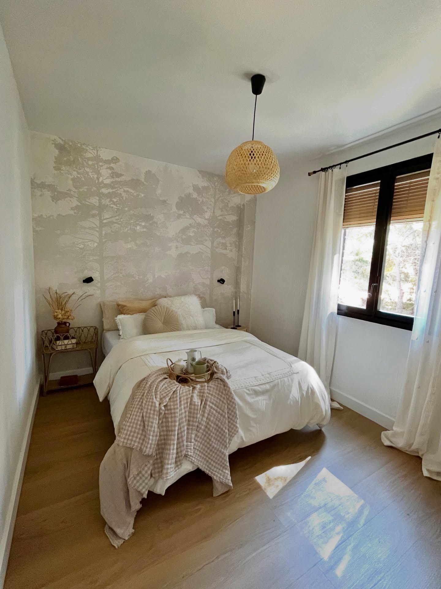 Una camera da letto accogliente con un letto bianco e un plaid beige a quadri. La parete dietro il letto ha una carta da parati grigio chiaro con alberi. Una lampada a sospensione rotonda in vimini è appesa e una finestra con tende bianche si trova sulla destra.