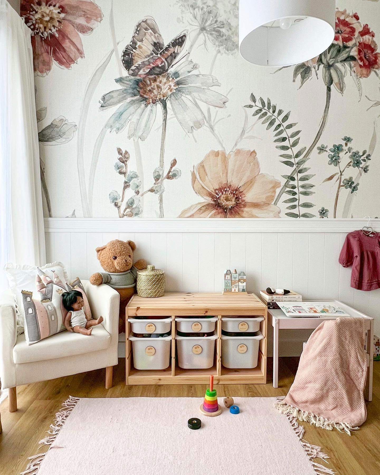 Child's room with floral wallpaper, white chair, wooden storage unit with bins, pink rug, and toys.