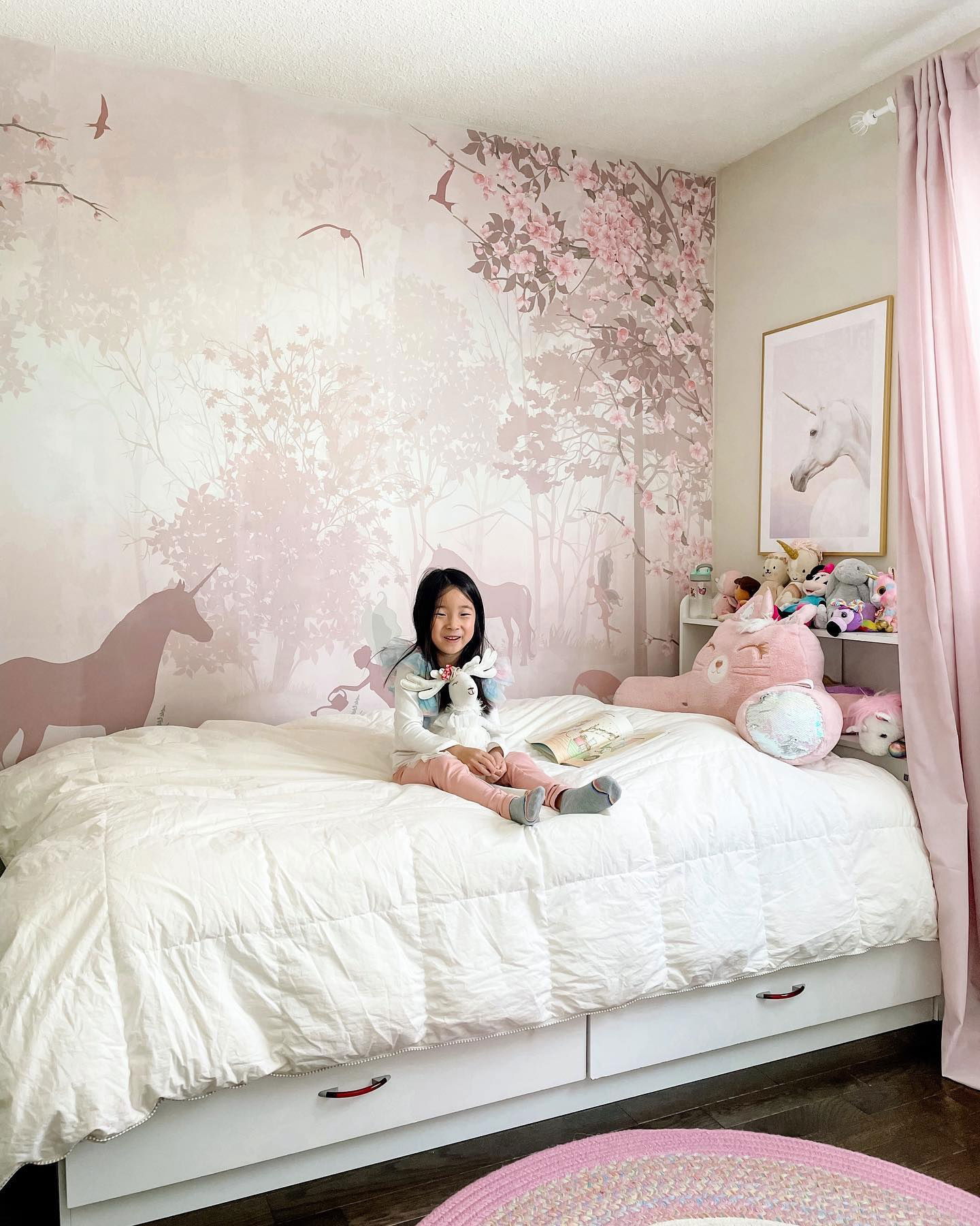A girl sits on a bed in a unicorn-themed room. The wall has a forest mural with unicorns and fairies. A unicorn print hangs near pink curtains.
