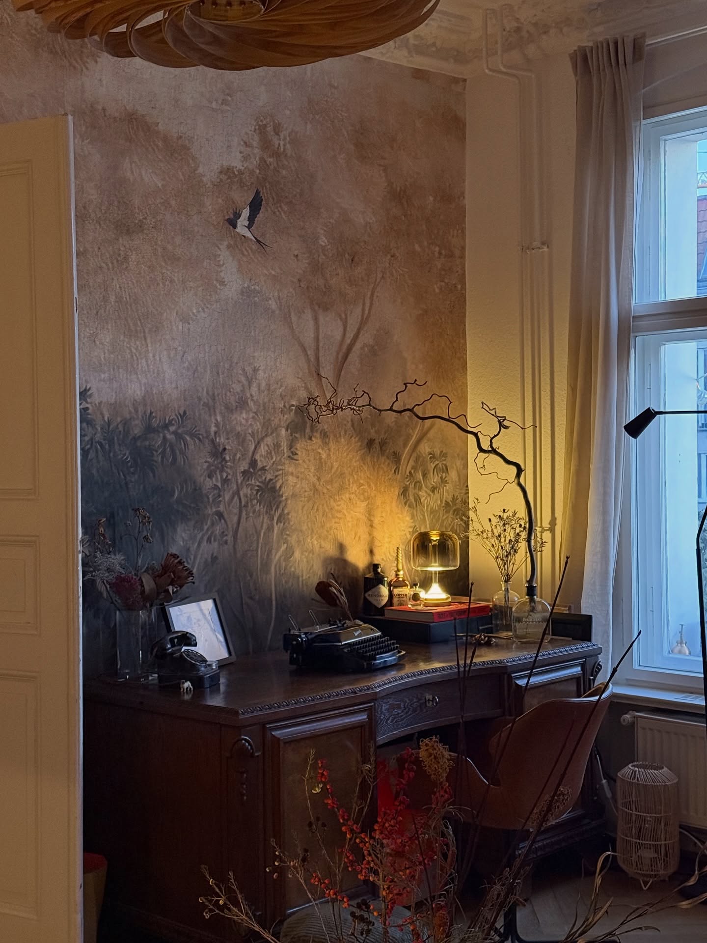 Een donkerhouten bureau met een vintage zwarte typemachine, lamp en gedroogde takken staat tegen een gedempte bosmuurschildering met een zwart-witte vliegende vogel. Rechts staat een raam.