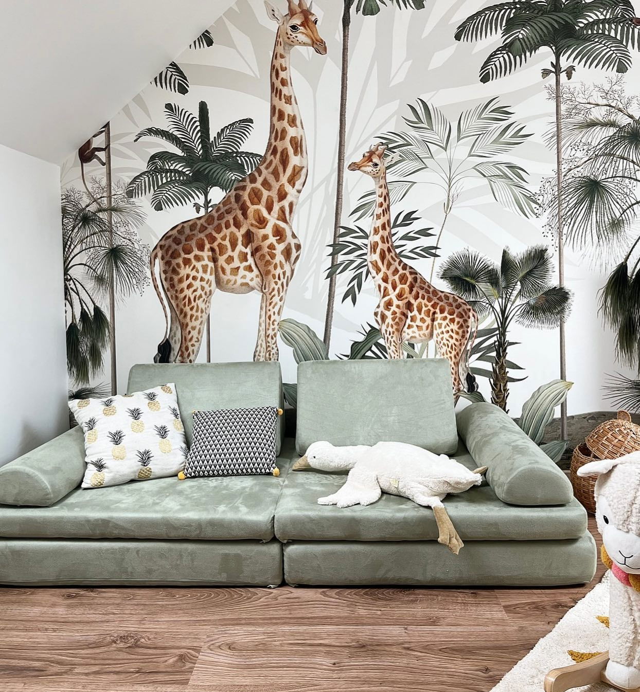 Intérieur avec canapé modulable vert sauge, coussins et animaux en peluche. Papier peint girafe et palmier.