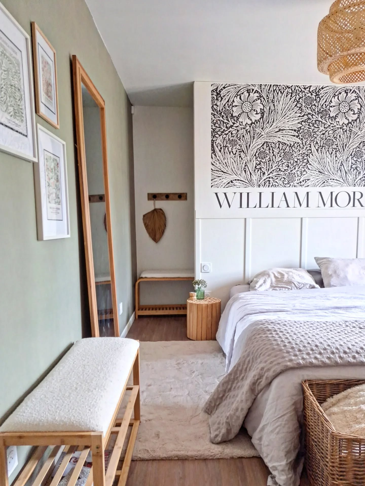 Makuuhuone, jossa valkoiset vuodevaatteet ja harmaa peitto sängyllä. Valkoisen paneelipäädyn yläpuolella on suuri mustavalkoinen kasvitieteellinen printti, jossa on teksti "WILLIAM MORRIS". Vaaleanvihreät seinät ja puiset huonekalut.