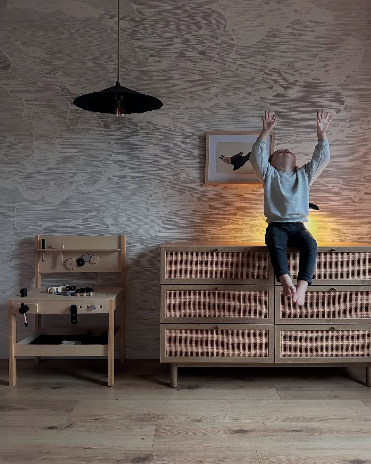 Child sitting on a wooden dresser with woven drawers in a nursery with cloud wallpaper.
