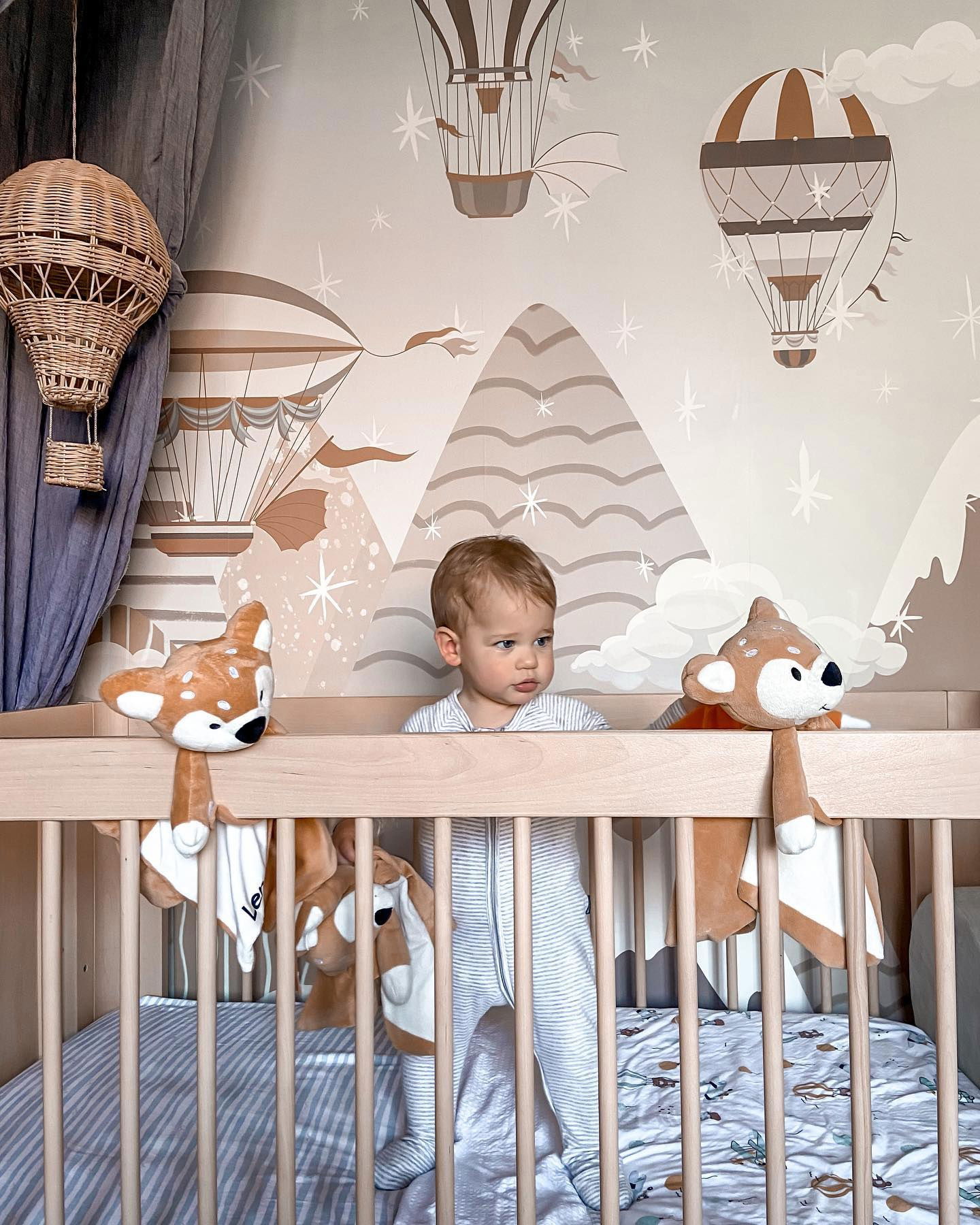 Baby im Kinderbett mit Hirschplüschtieren, Heißluftballon-Tapete und Korbballonlampe.