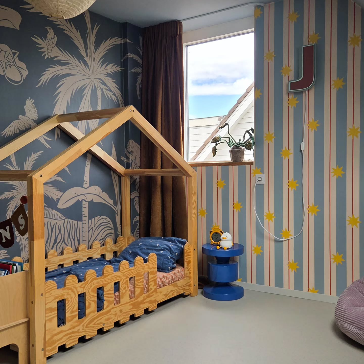 A wooden house-shaped bed with blue bedding in a room with tropical and striped wallpaper.
