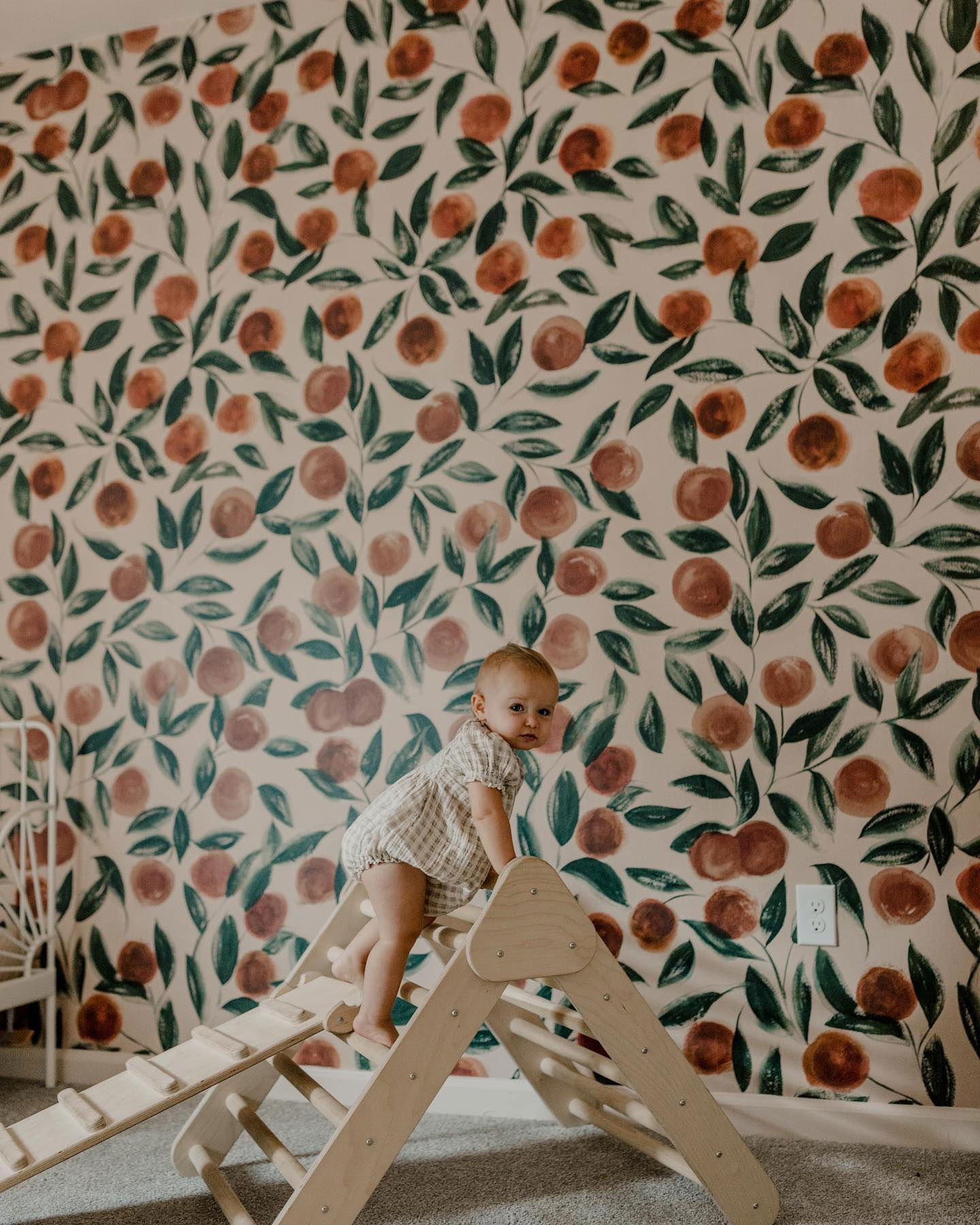 Baby climbing a wooden Pikler triangle in a room with orange fruit wallpaper.