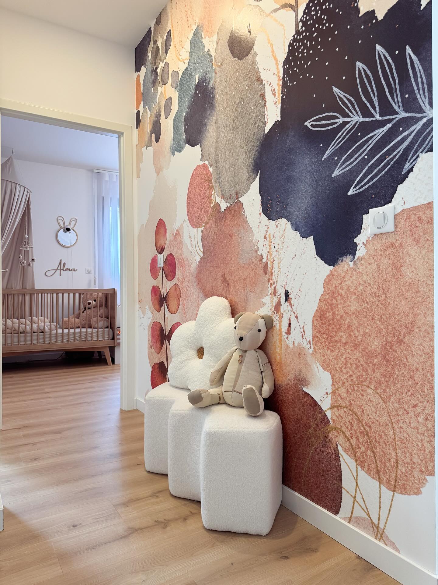 Hallway with abstract watercolor wallpaper, teddy bear on white ottoman, and nursery view.