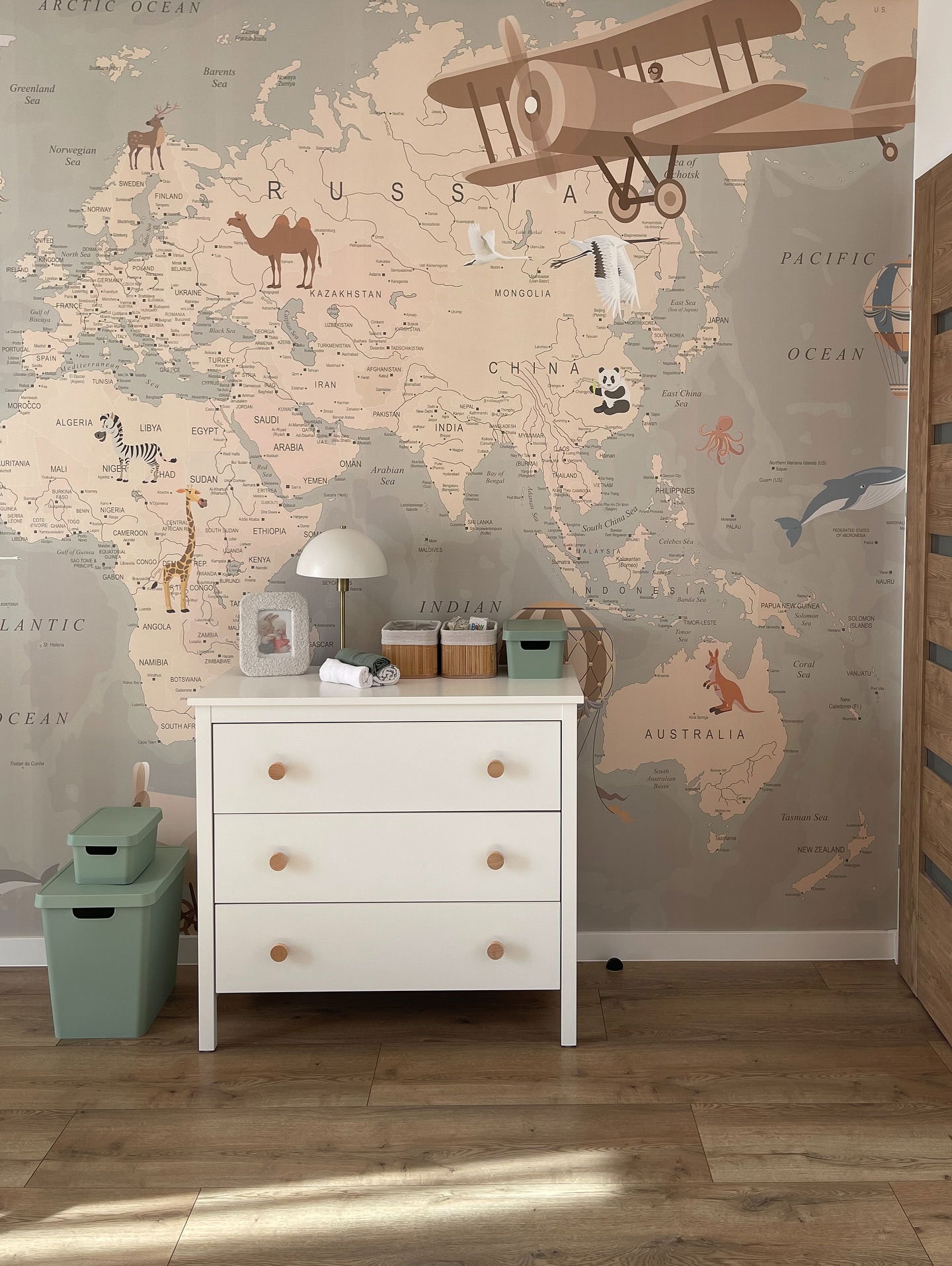 Stanza dei bambini con comò bianco, pomelli in legno e carta da parati con mappamondo.